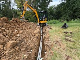 Excavator digging a trench beside a large dirt pile in a grassy field