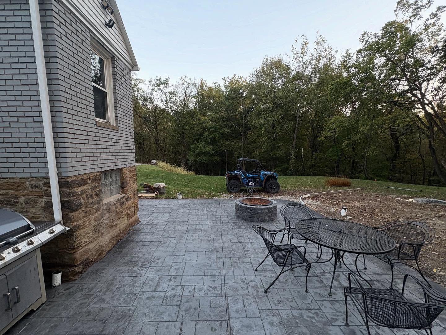 Stone patio with metal chairs and a fire pit beside a house, overlooking a grassy yard and trees.