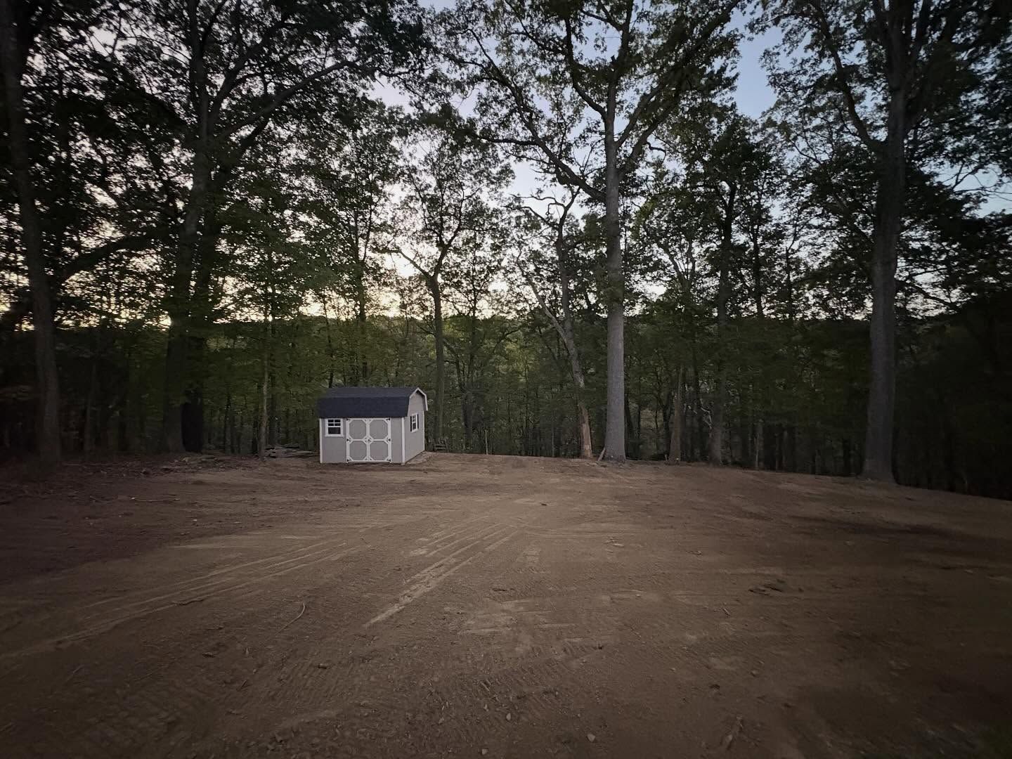 Small white shed in a cleared dirt area, surrounded by tall trees at dusk.