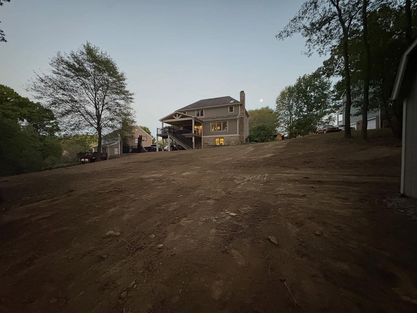 Dirt slope leading to a lit house at dusk, with trees framing the yard.