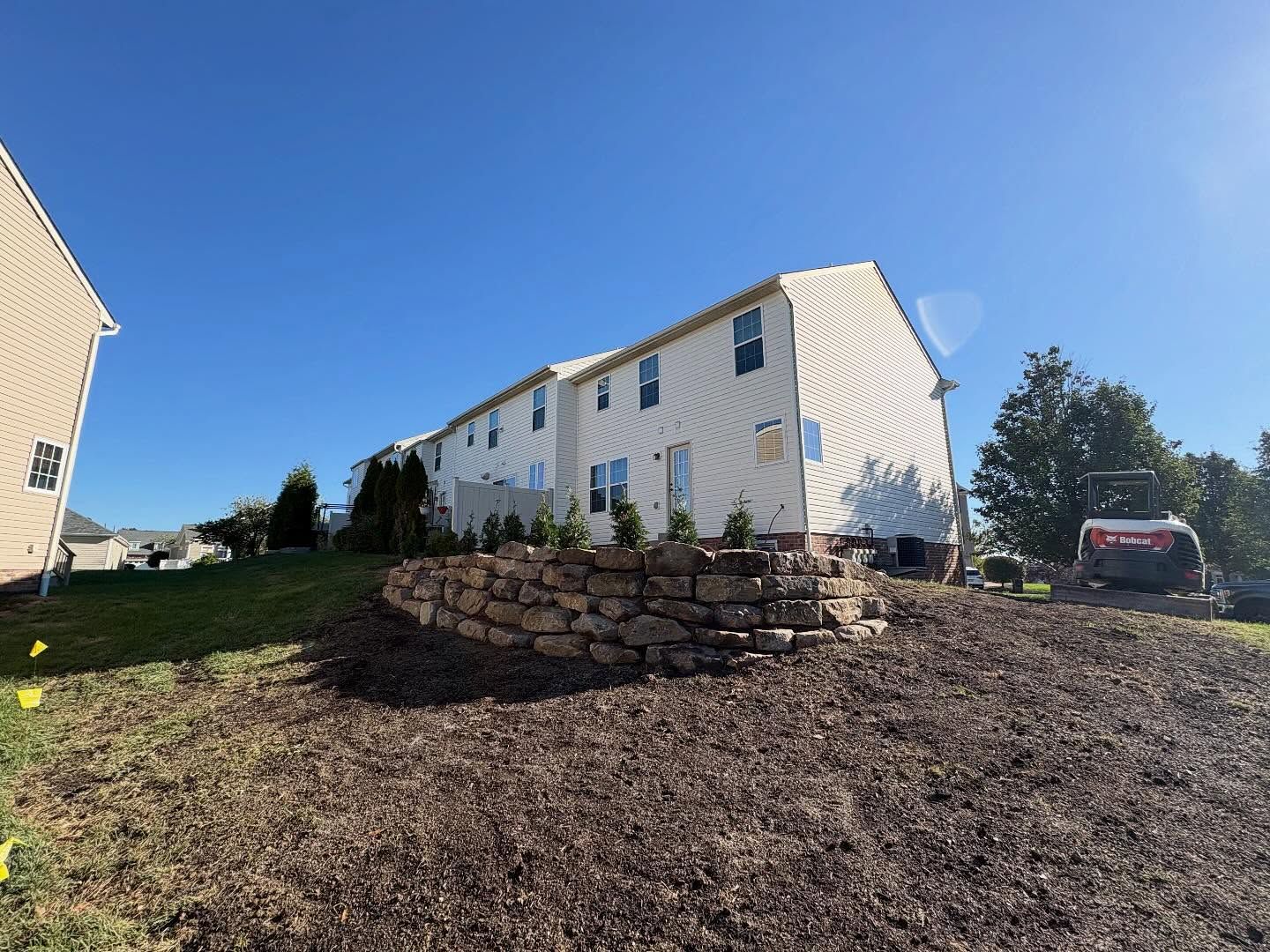White house with blue-trimmed windows on a sloped yard, stone retaining wall, and parked red car under a blue sky
