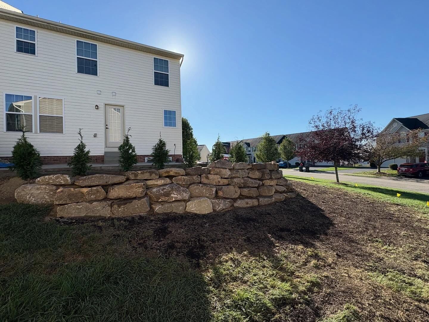 White house beside a stone retaining wall under a bright blue sky, with a grassy yard and trees nearby