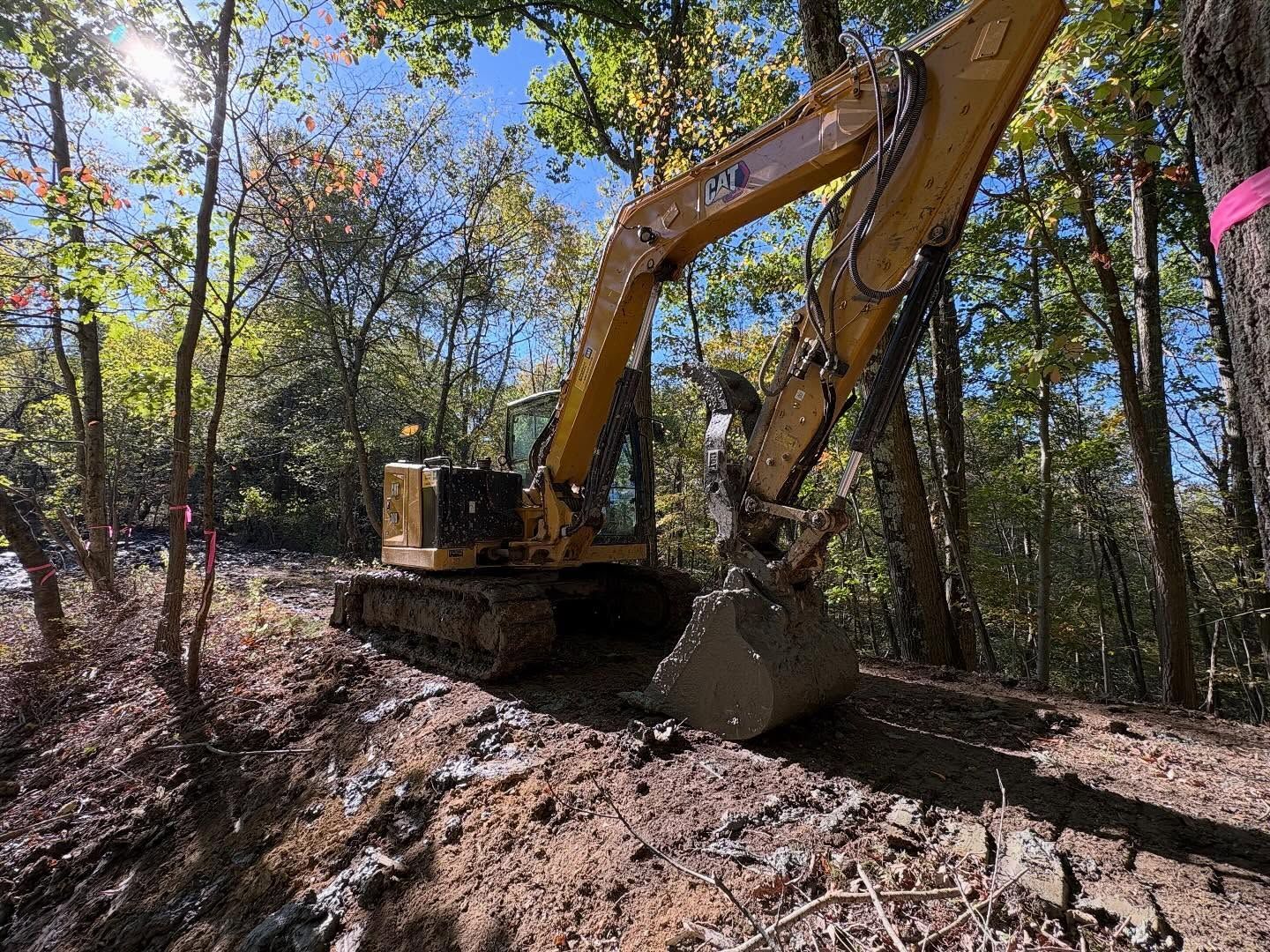 Yellow excavator working on a muddy wooded trail under trees in autumn