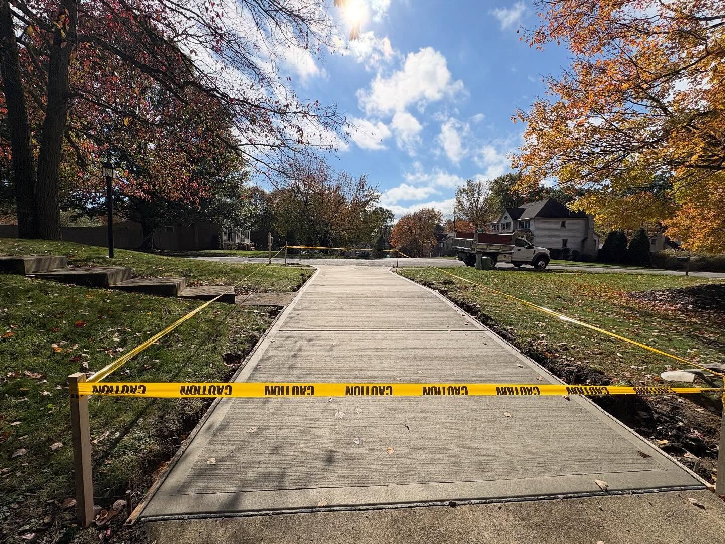 Yellow caution tape blocks a concrete park path lined with autumn trees and houses in the background