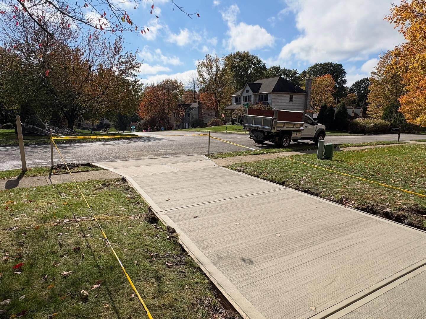 Concrete sidewalk ramp leading to a residential street, with a truck and autumn trees nearby.