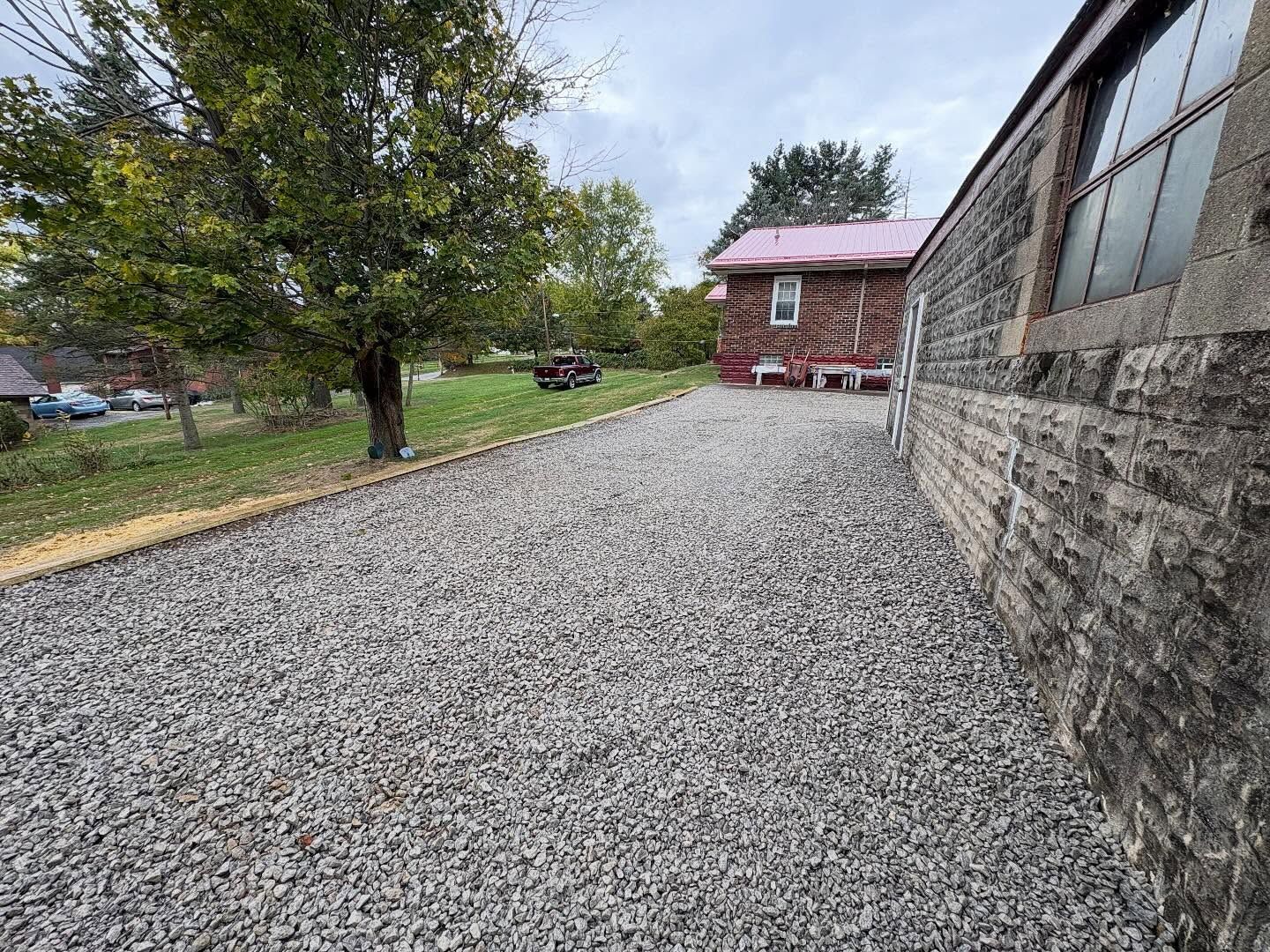 Gravel driveway beside a stone wall, leading to a red brick building and trees on a cloudy day