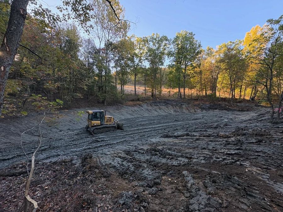 Bulldozer clearing a muddy field beside autumn trees under a clear sky.