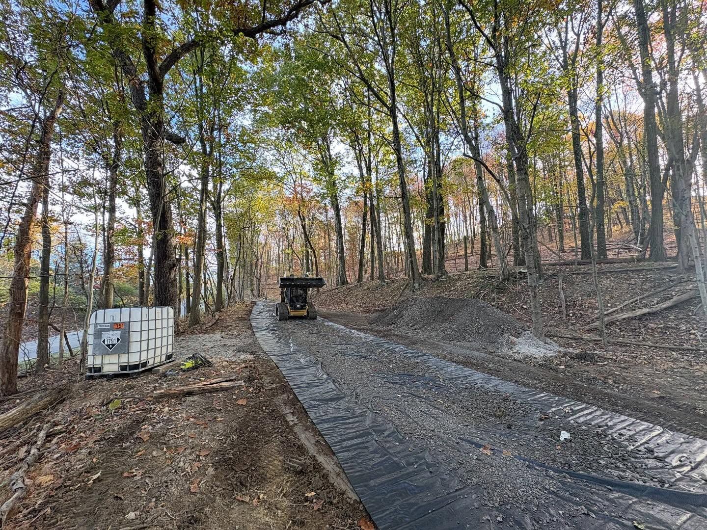 Gravel driveway under construction in a wooded area, with a dump truck and white container nearby.