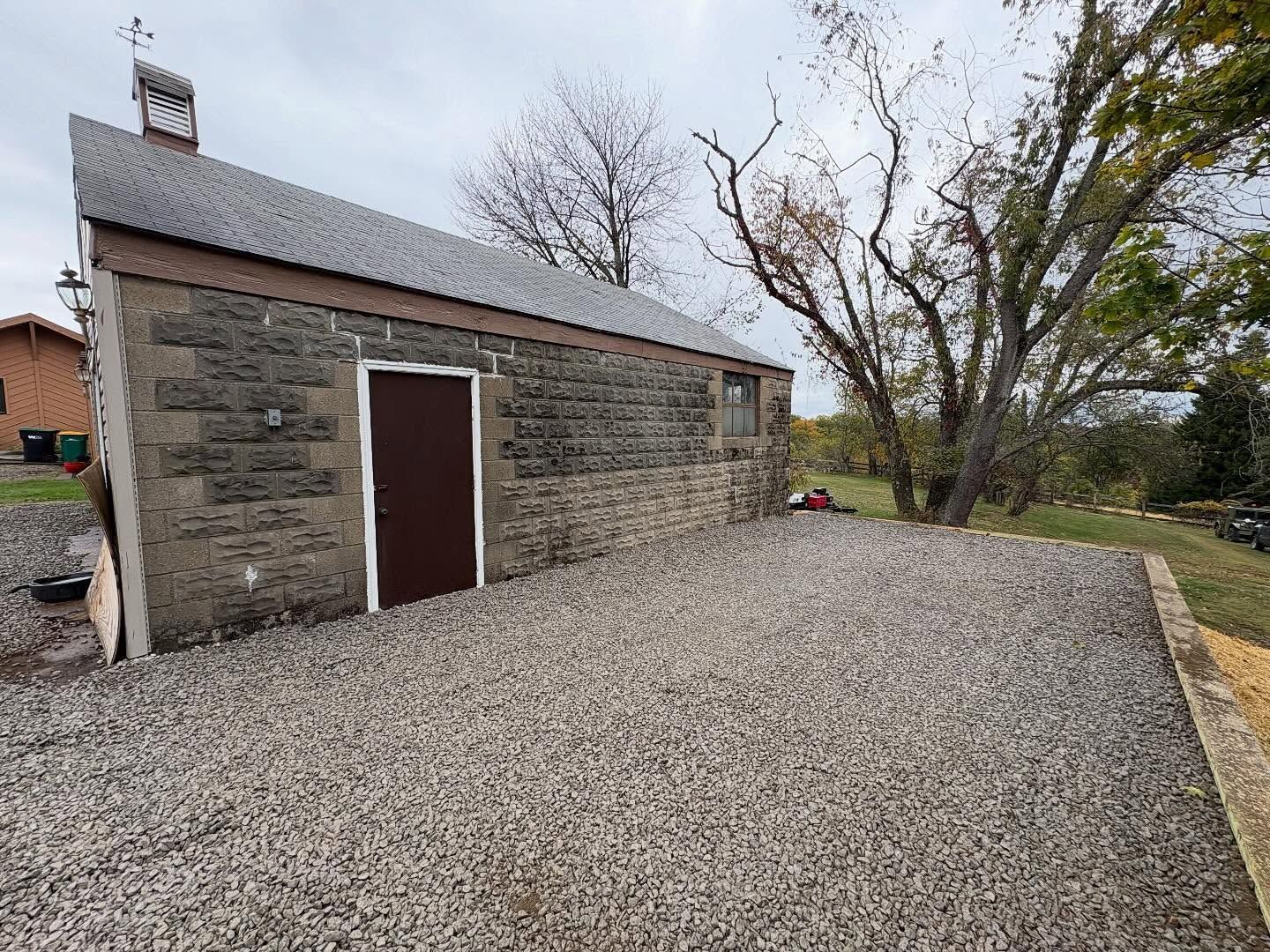 Stone shed with brown door on a gravel lot beside leafless trees