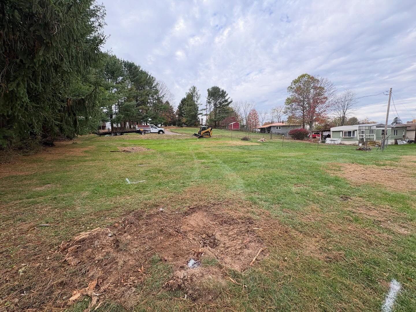 Grassy vacant lot with dirt patches, trees on the left, and houses in the background under a cloudy sky