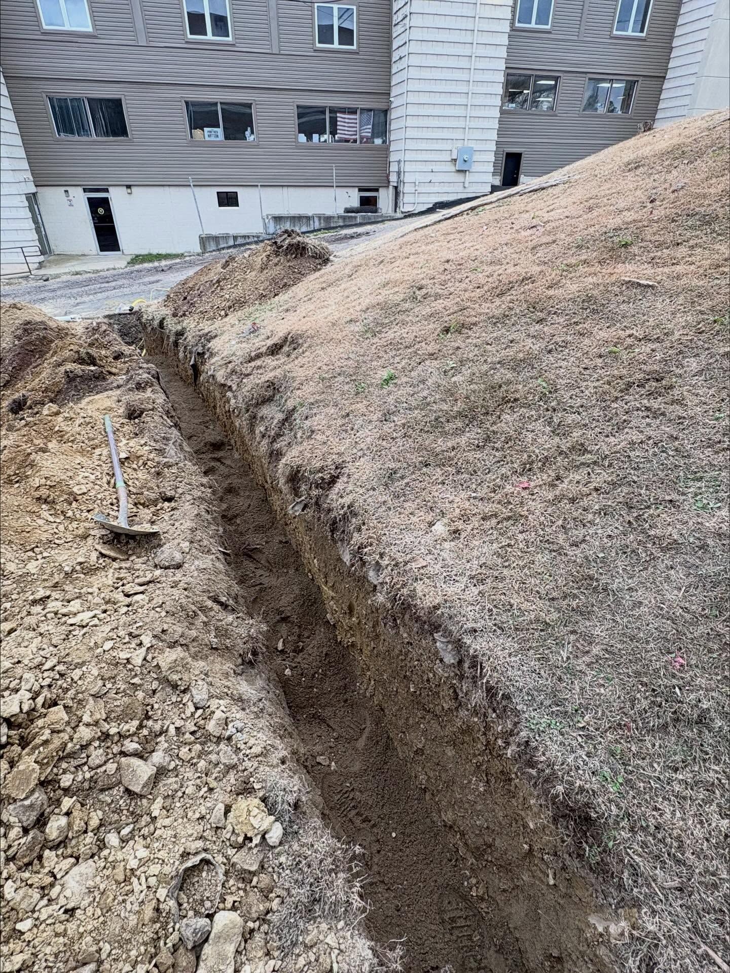 Trench cut through a grassy dirt slope near apartment buildings with a shovel in the foreground