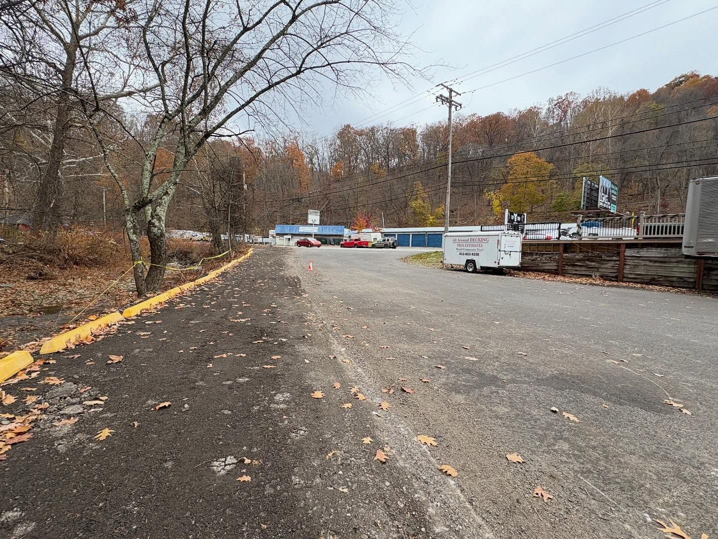 Empty road leading to a closed gate at a wooded entrance, with fallen leaves and parked vehicles beyond