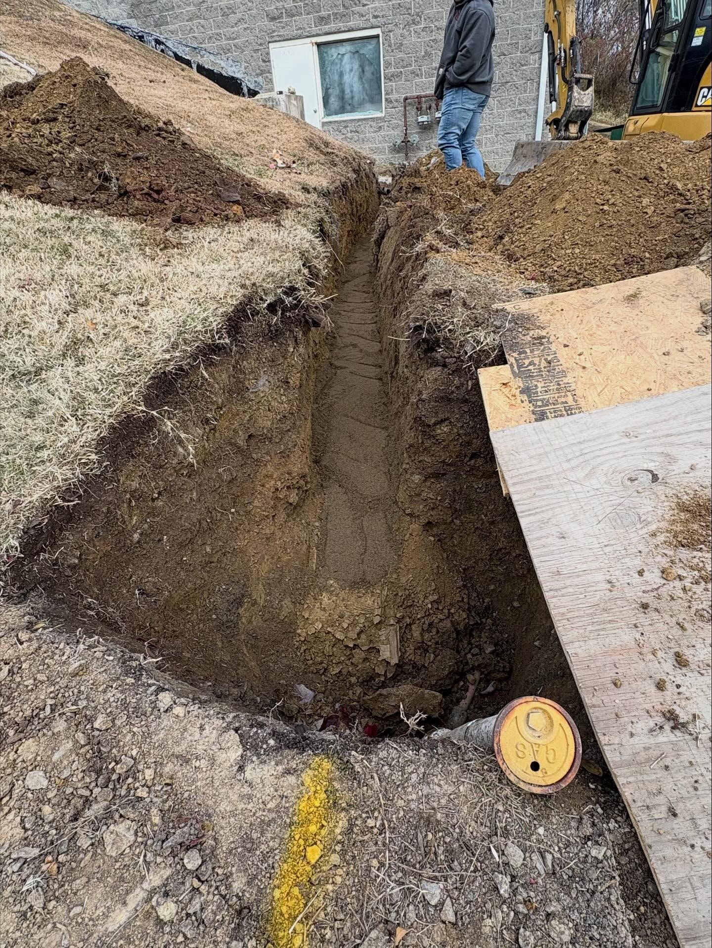 Deep trench dug beside a building, with soil piles and a measuring tape on the ground.