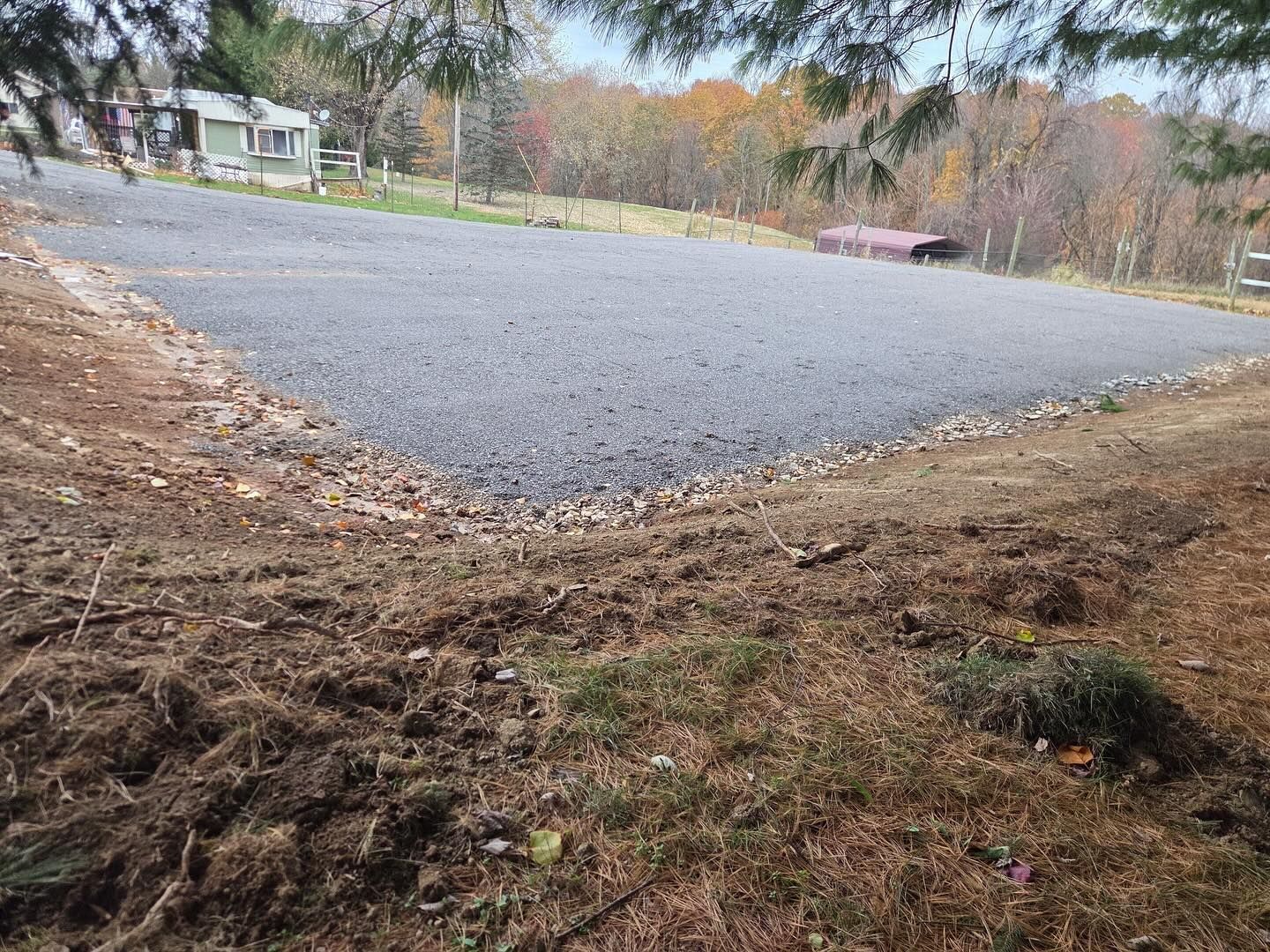 Gravel parking area beside a wooded yard, with a house in the background and a bench near the trees.