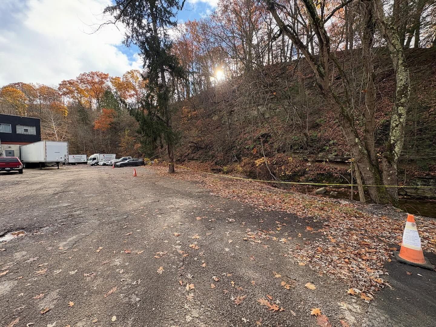 Gravel parking lot beside a wooded hillside with parked trucks and an orange traffic cone