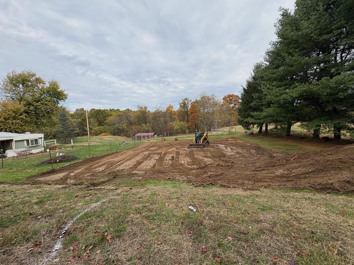 Bulldozer grading a muddy lot beside grassy trees under a cloudy sky