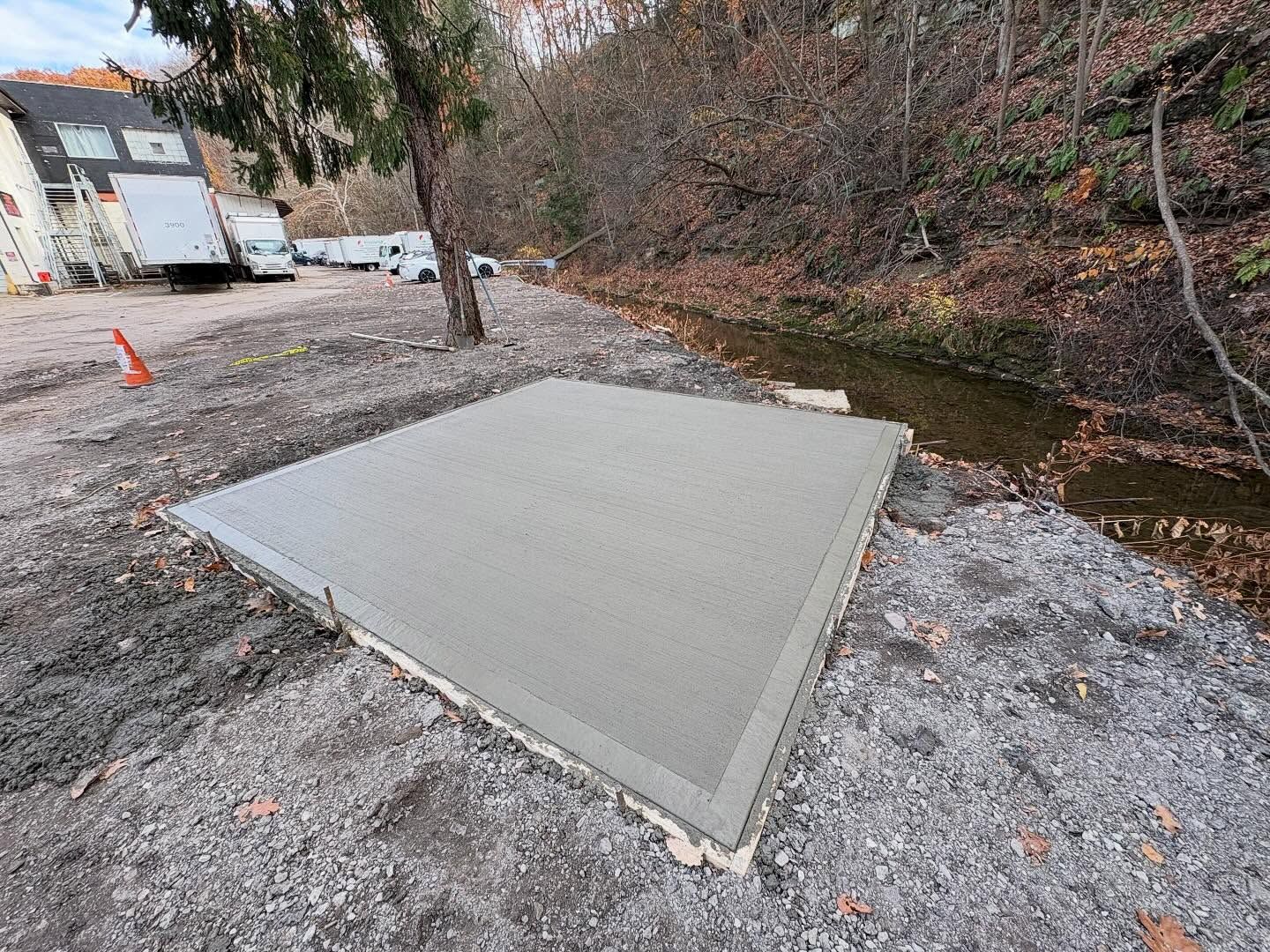 Freshly poured concrete slab beside a creek, with gravel, trees, and construction equipment nearby