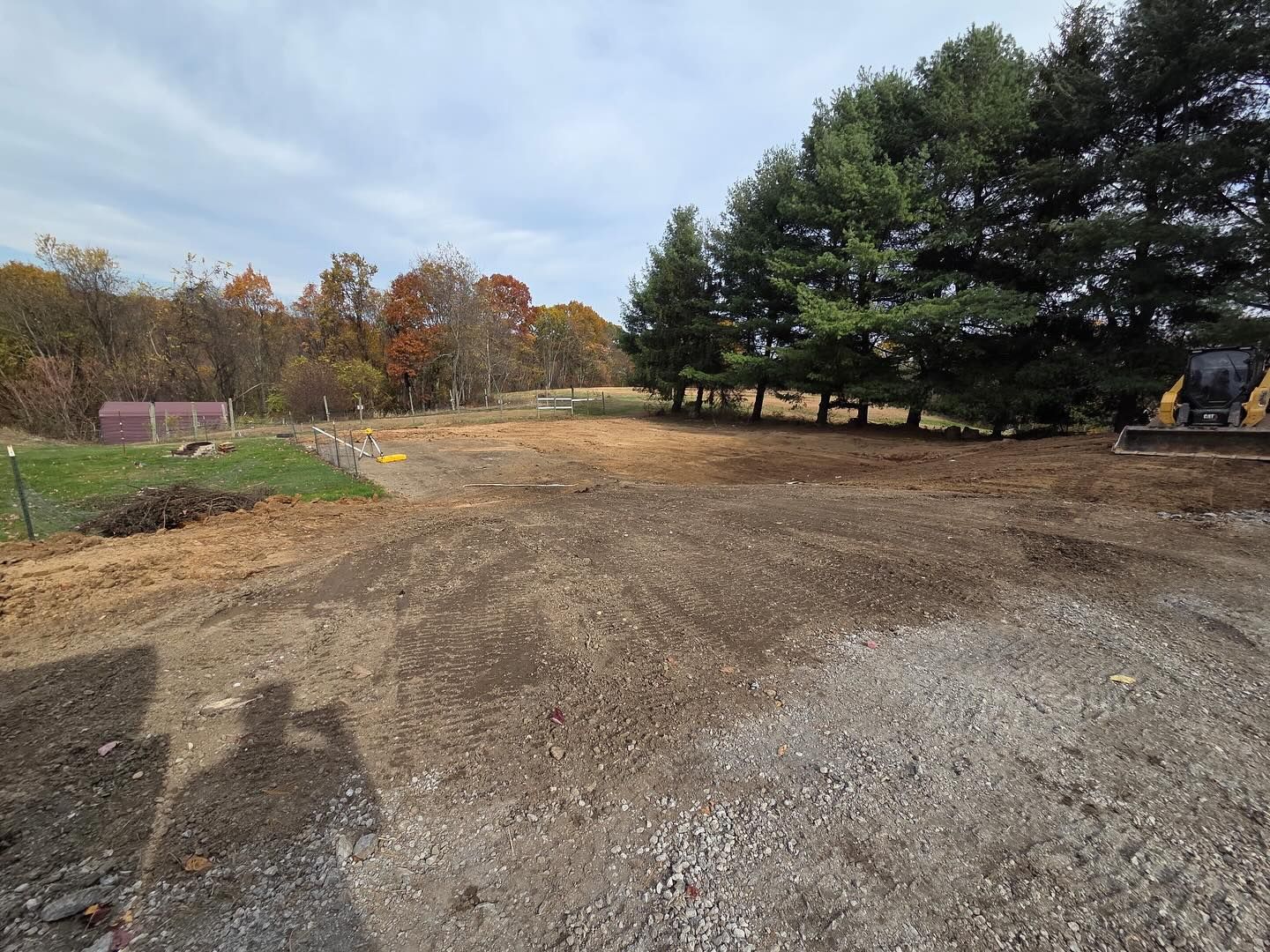 Gravel lot beside trees and a small red barn under an autumn sky, with heavy equipment at right