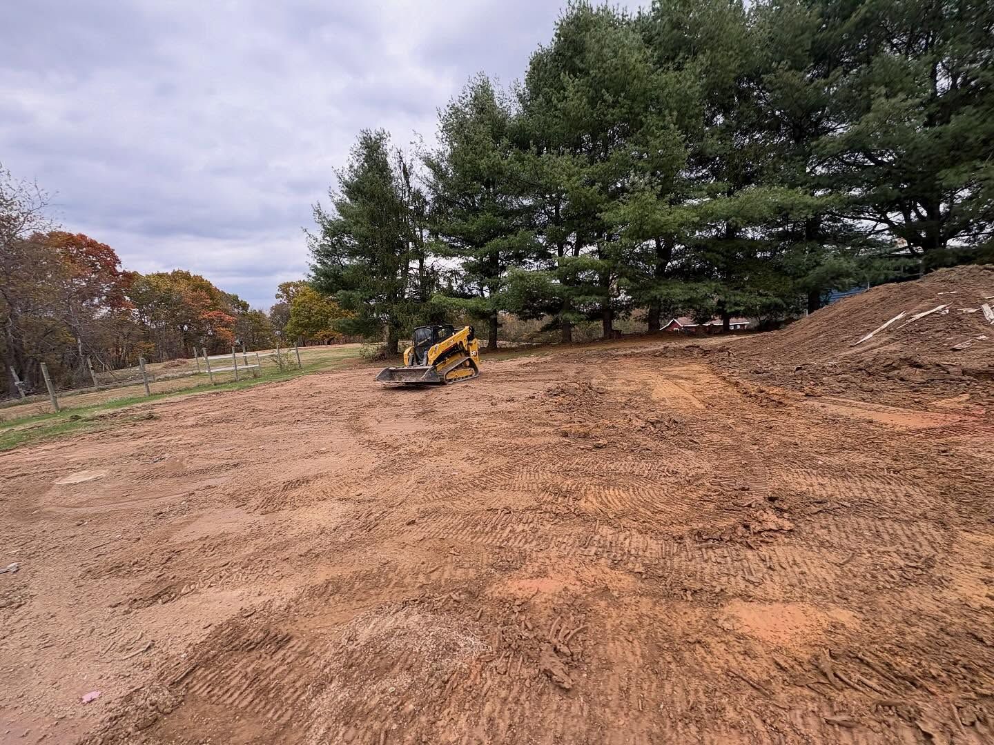 Yellow excavator on a cleared dirt lot beside pine trees under a cloudy sky
