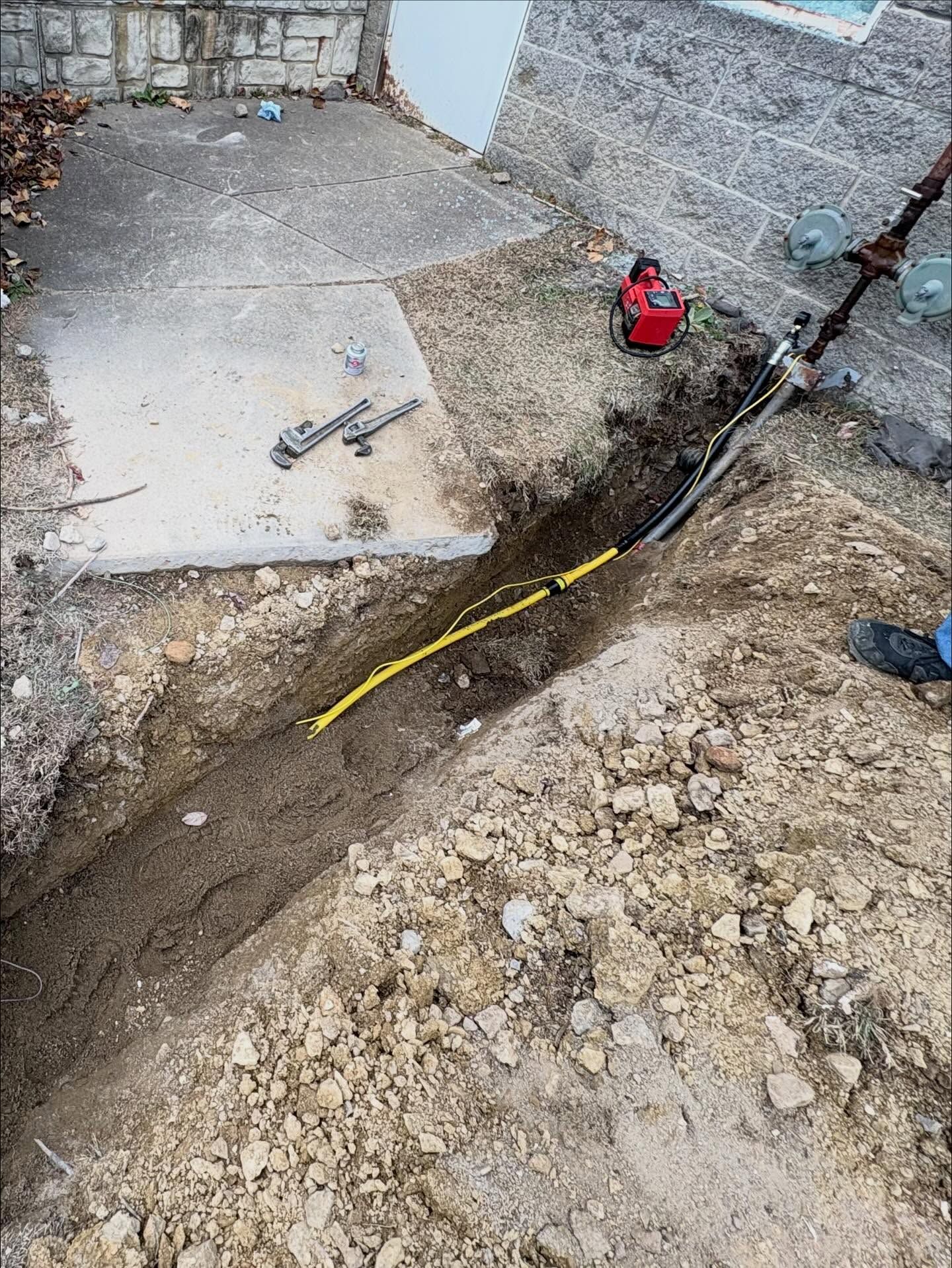 Trench dug beside a concrete slab at a construction site, with a red tool and yellow hose on the ground.