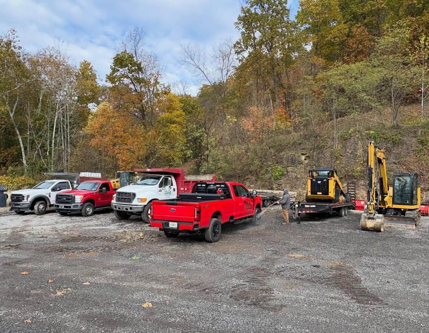 Parked pickup trucks and small construction equipment on a gravel lot beside autumn trees