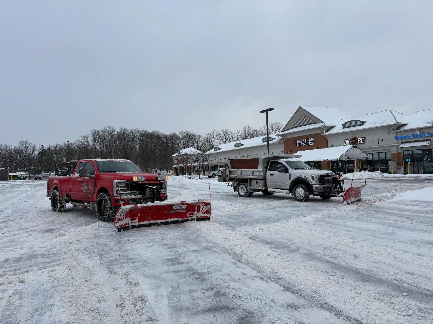 Red and white snowplow trucks clearing a snowy parking lot by a building