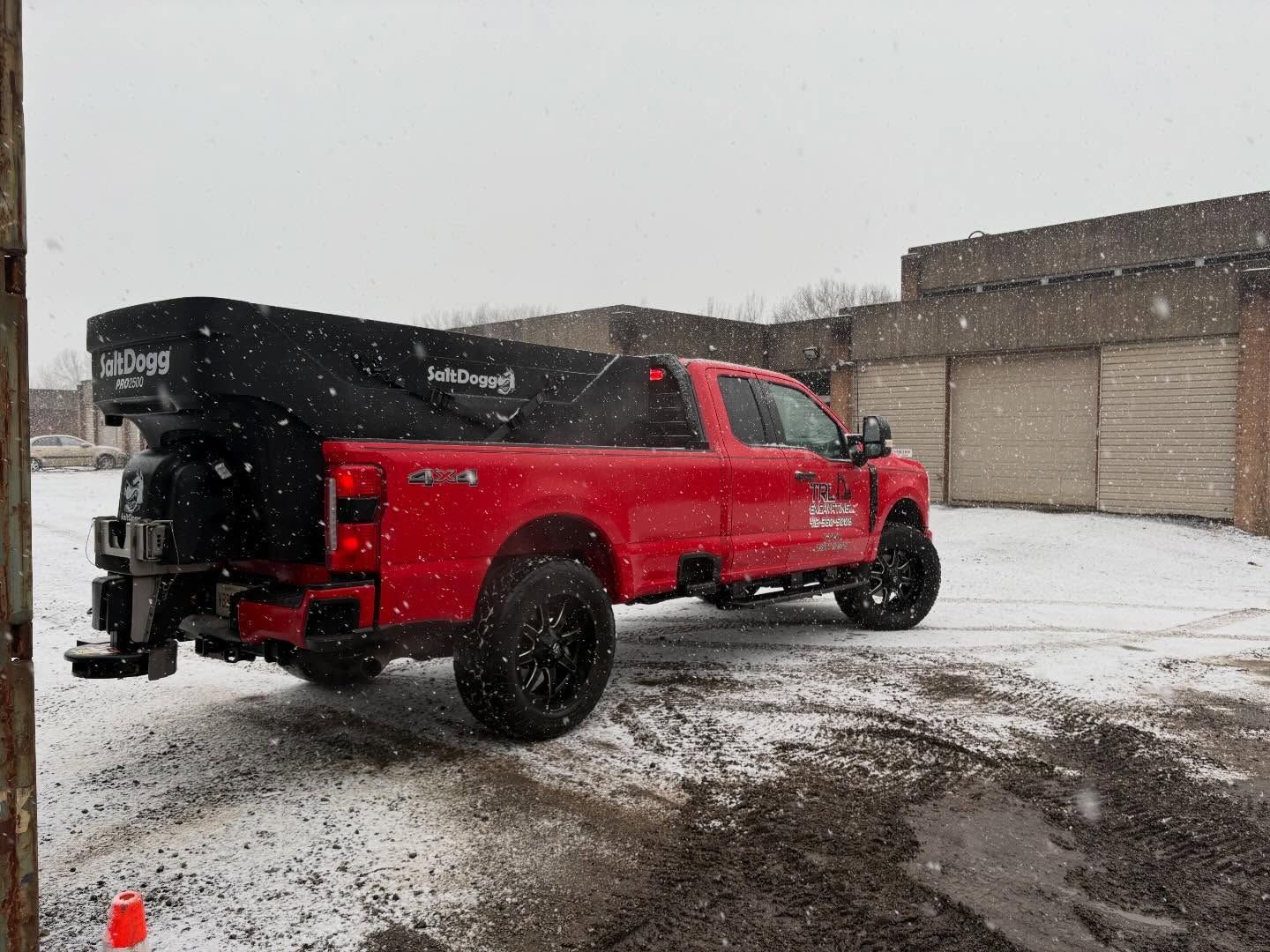 Red pickup truck with black canopy parked in a snowy lot near industrial buildings.