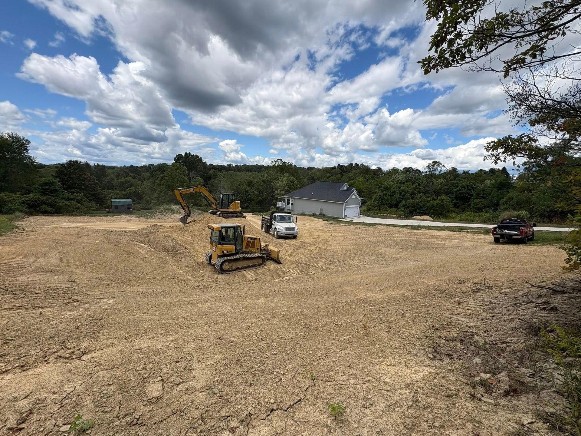 Cleared dirt lot with excavator and truck near a white house under a cloudy sky