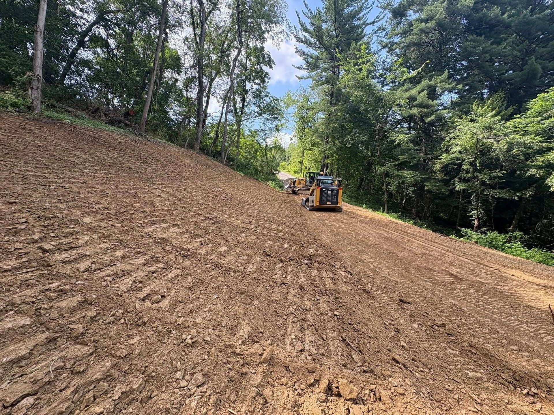 Tractor grading a dirt hillside beside trees under a blue sky