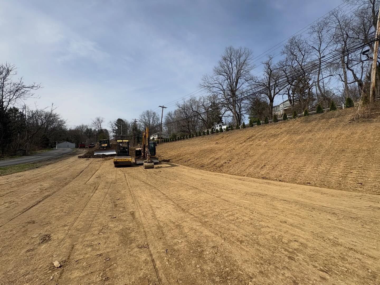 Construction equipment grading a dirt roadway beside a wooded hillside under a cloudy sky
