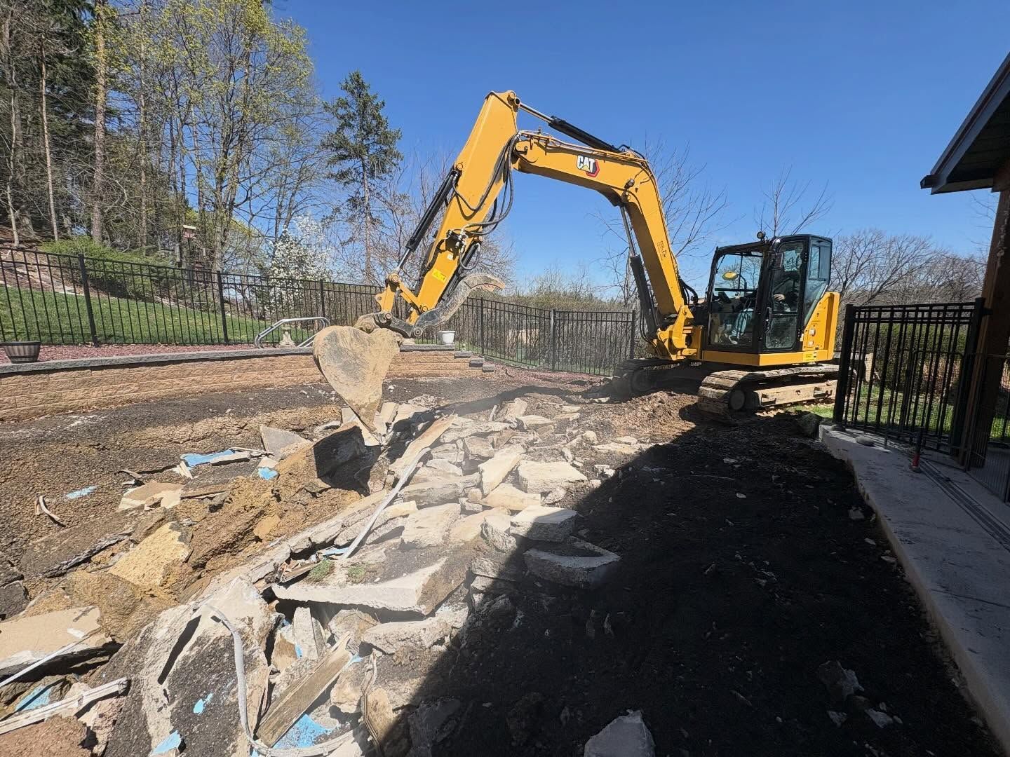 Yellow excavator demolishing concrete along a roadside beside a black fence under a blue sky