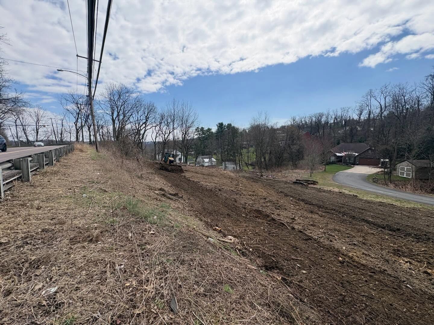 Dirt road cut along a wooded hillside under a partly cloudy sky, with a guardrail and houses nearby