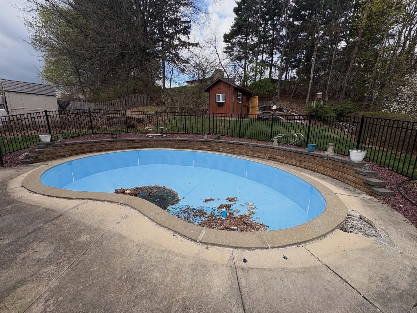 Empty drained backyard pool with blue lining and fallen leaves, surrounded by a fenced patio and trees.