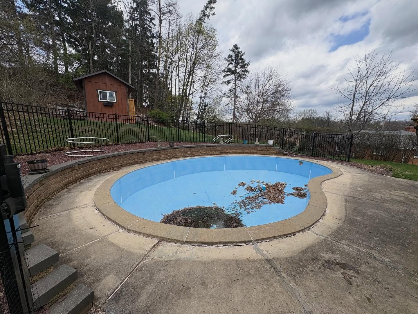 Empty blue swimming pool with leaves, beside a house and trees on a cloudy day