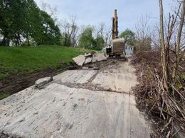 Construction excavator clearing a damaged concrete road beside a grassy embankment