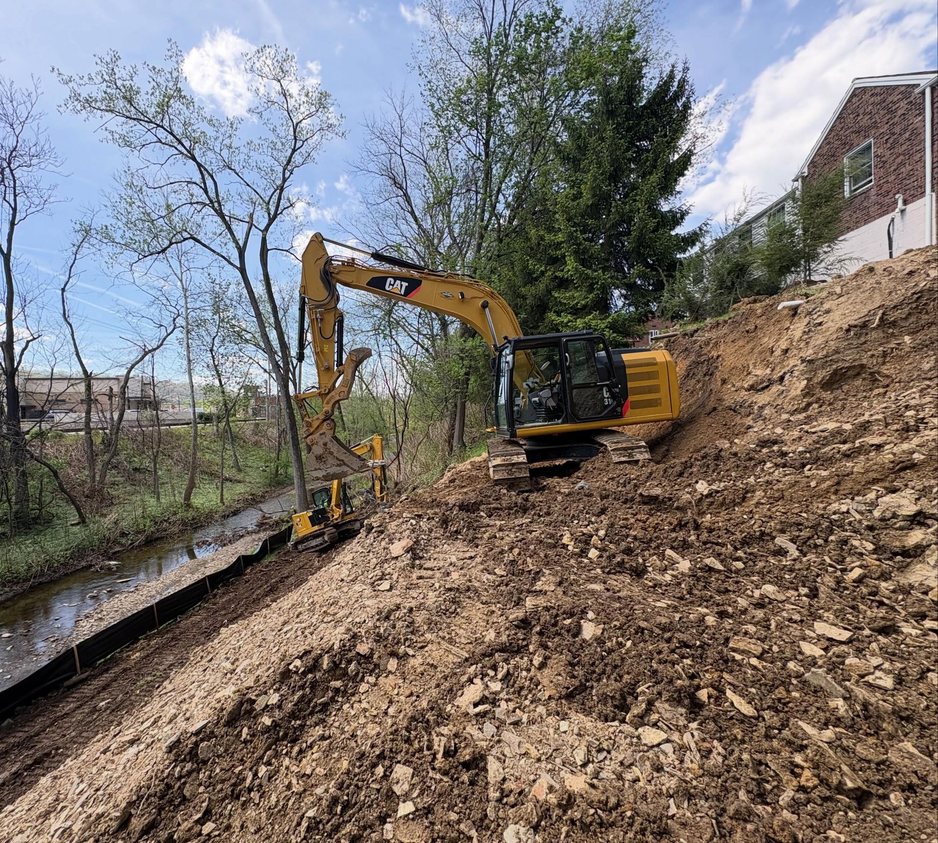 Yellow excavator working on a muddy hillside beside a stream and trees