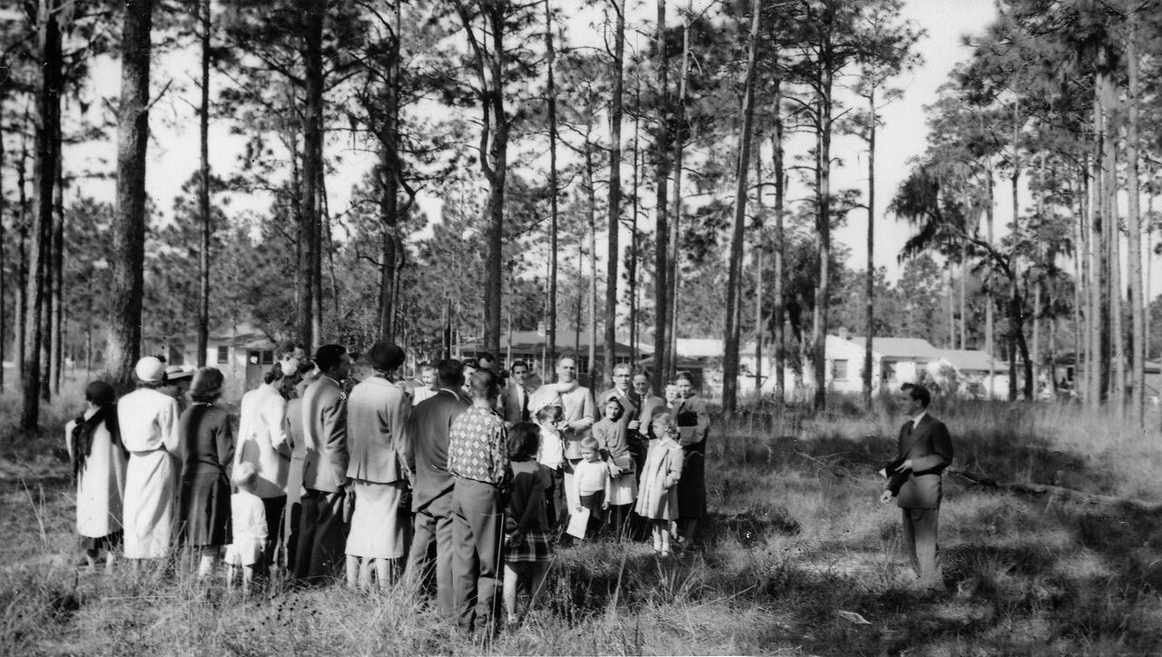 A group of people standing in a clearing in a forest, facing toward a structure in the background.