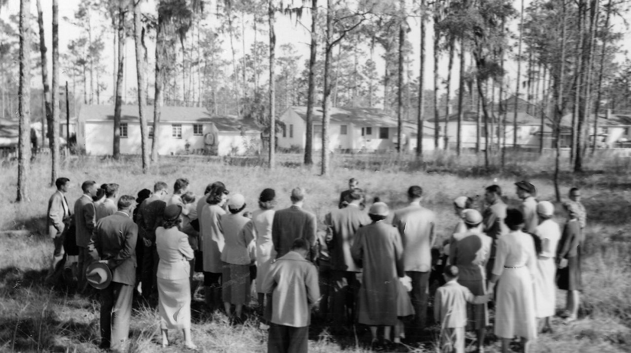 Group of people standing in a circle, facing a speaker, buildings in background, trees surrounding.