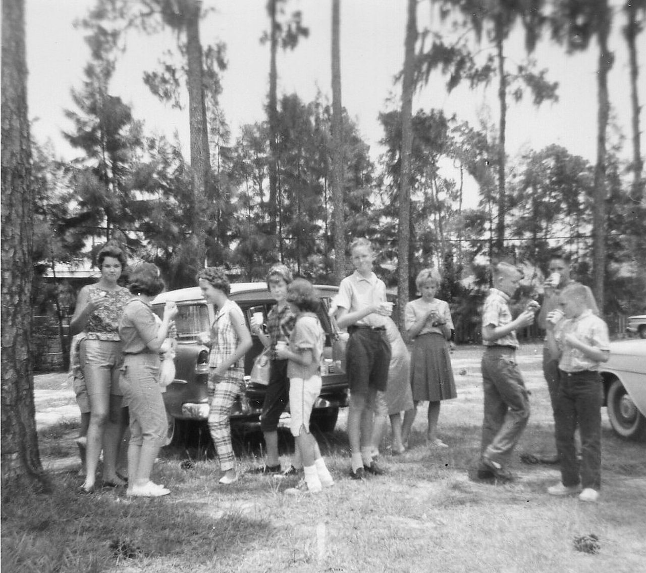 Group of people standing near cars in a grassy area, trees in the background. Black and white photo.