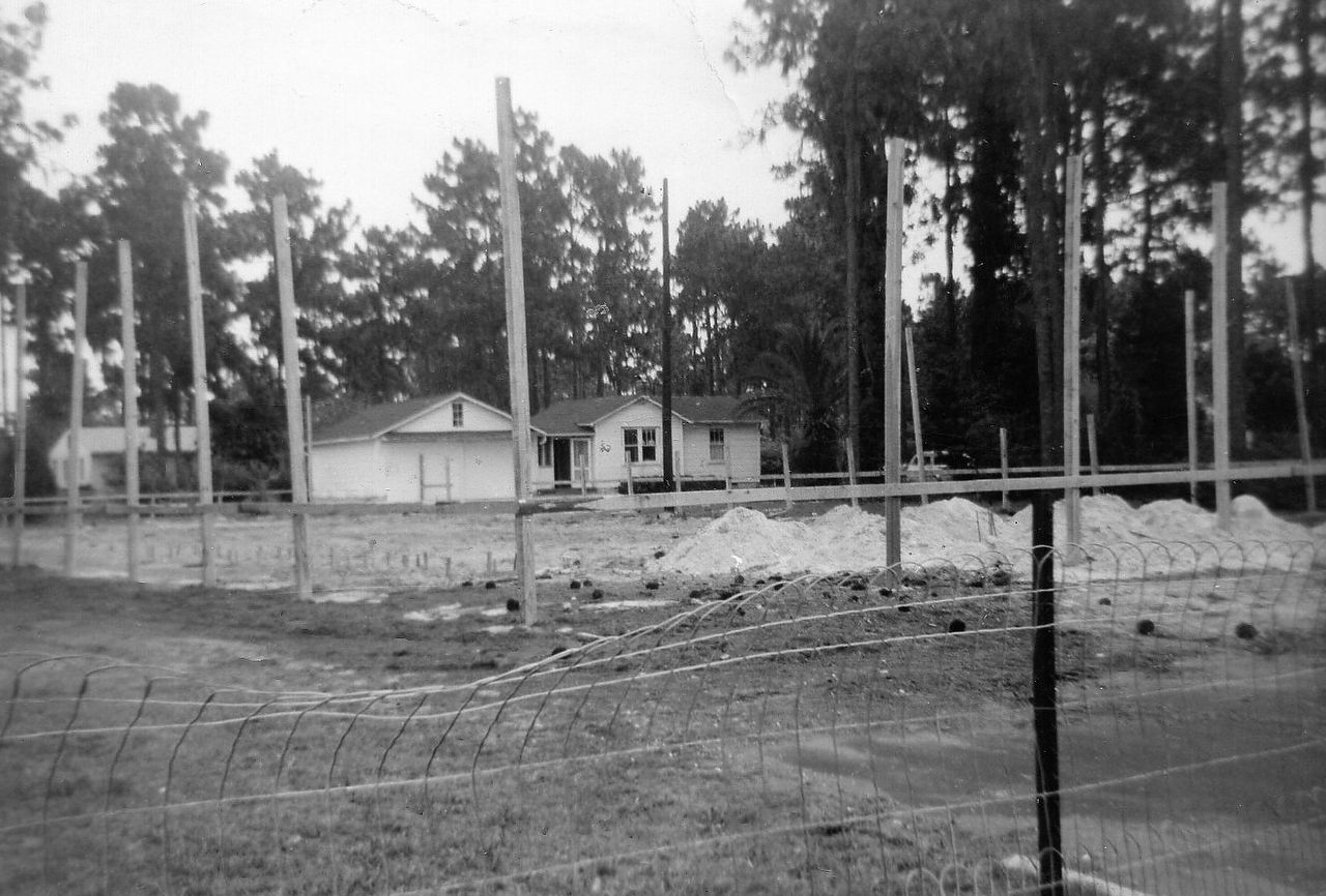 Black and white photo of a house under construction behind a wire fence and utility poles.