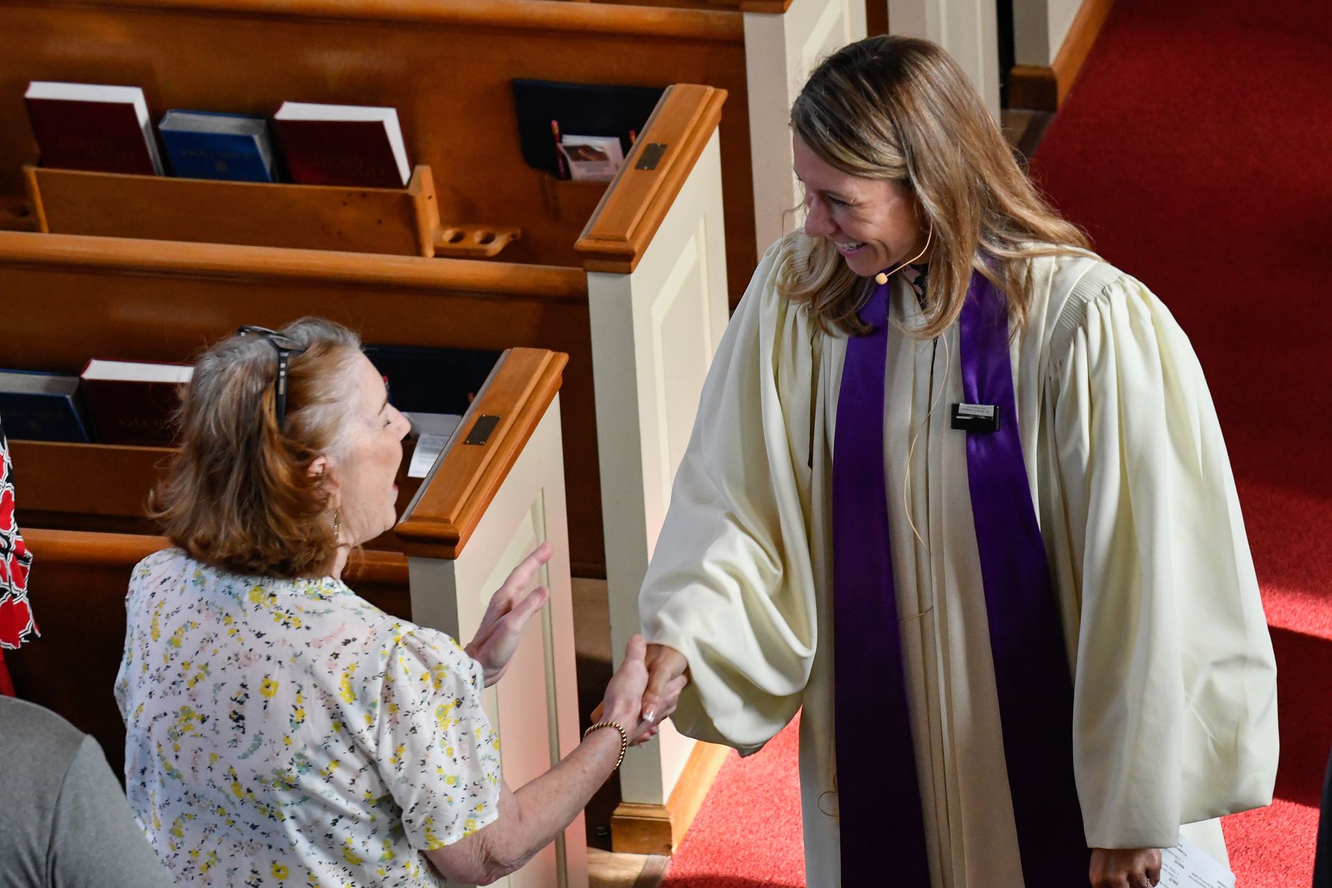 Clergy member in robe greets a woman in a church. Purple stole, wooden pews, red carpet.