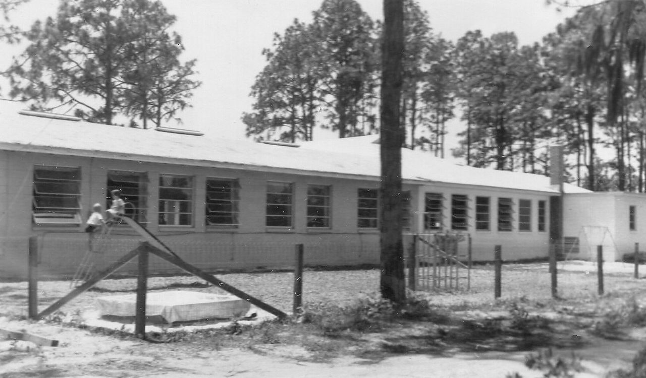 Long, single-story building with many windows, fence, and a playground, trees in the background.
