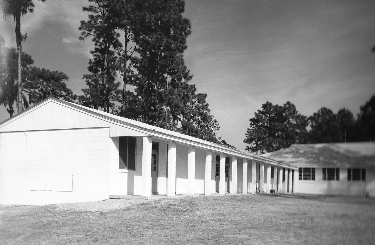 White, single-story building with columns and windows; trees and sky in the background.