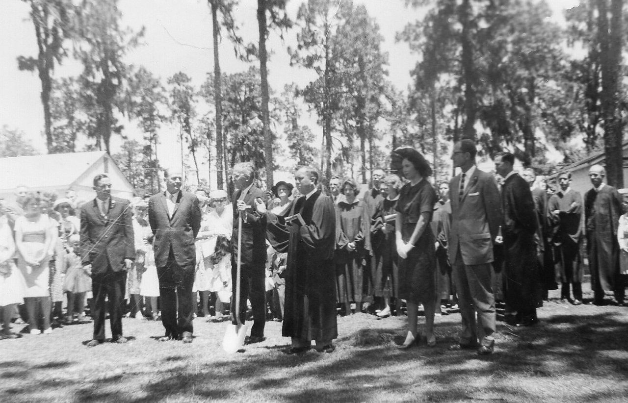 Group of people standing outdoors, some holding objects, under trees. Sunlight casts shadows.