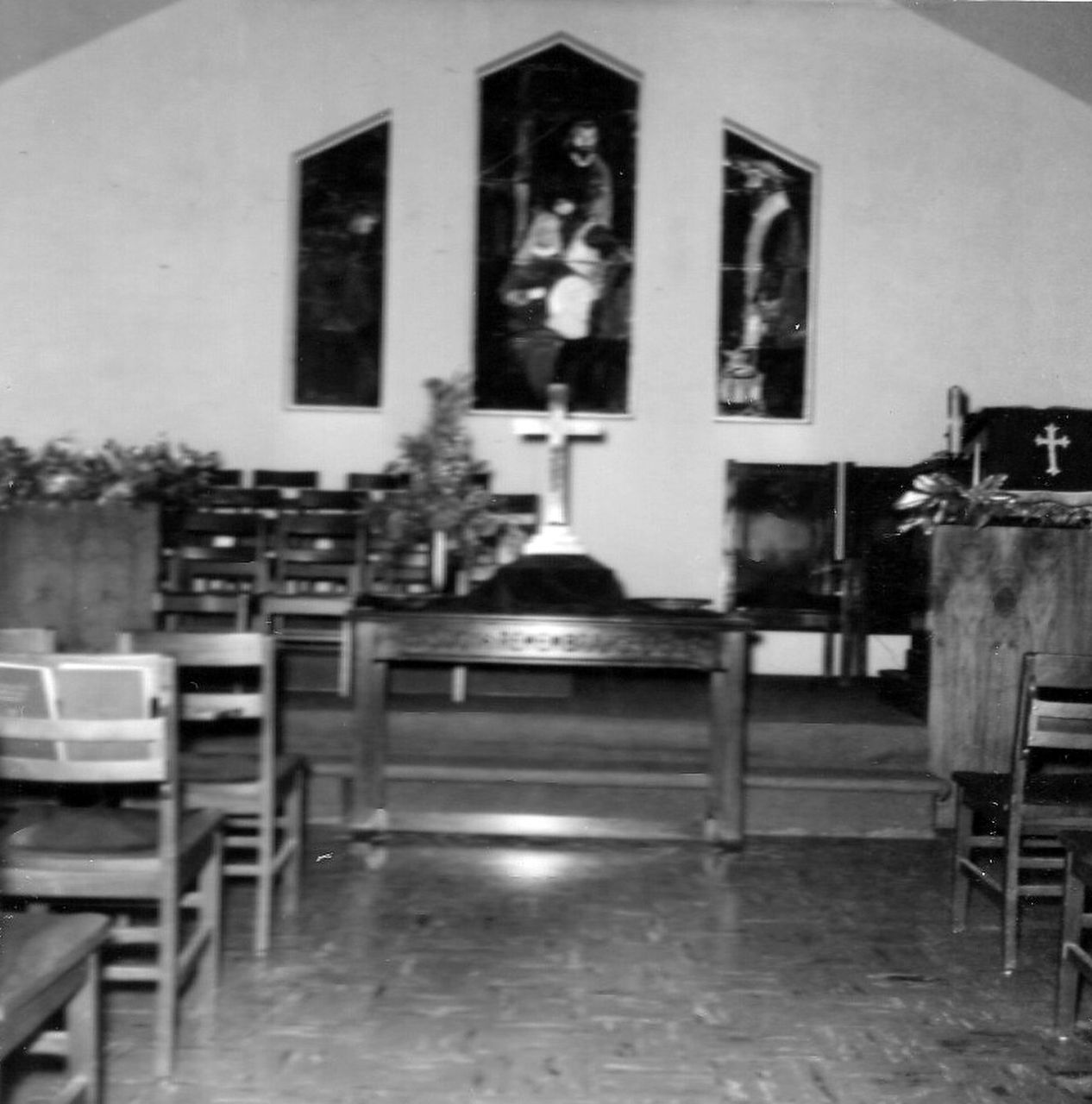 Interior of a church with wooden furniture, crucifix, and paintings on the wall. Black and white.