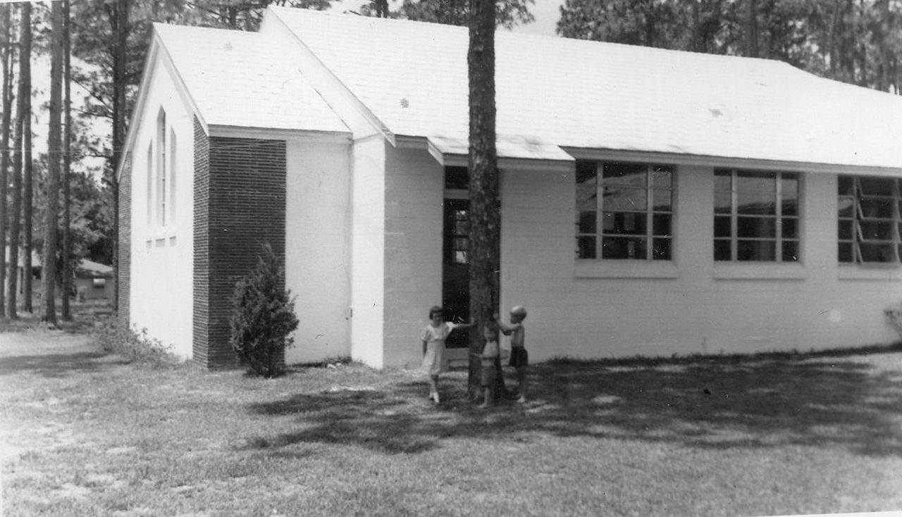 White, one-story building with a sloped roof and several windows. A tree stands in front, two people are in the doorway.