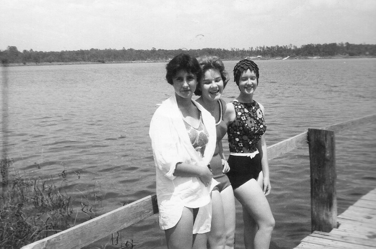 Three women in swimwear on a wooden dock, lake in background.