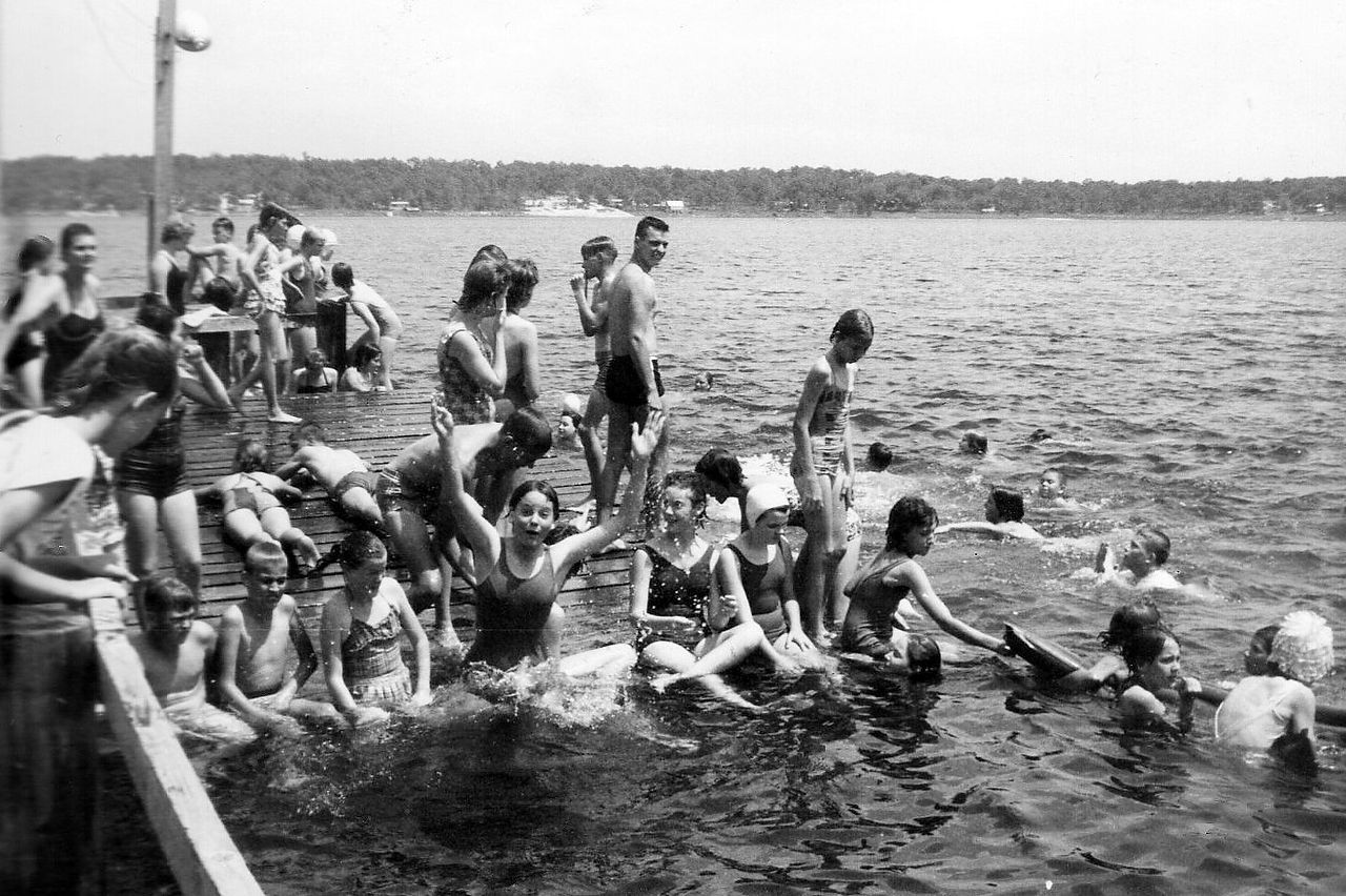 People swimming and on a pier in a lake. Many are in the water, some looking at the camera. Trees line the far shore.