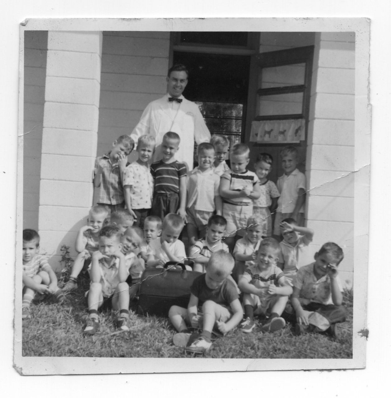 A group of children and a man in a bow tie pose outside a building, likely a school.