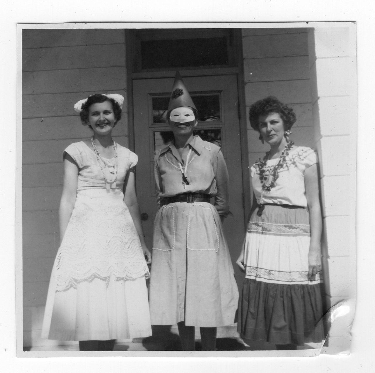 Three people in costume pose in front of a building with a door. One wears a mask and hat.